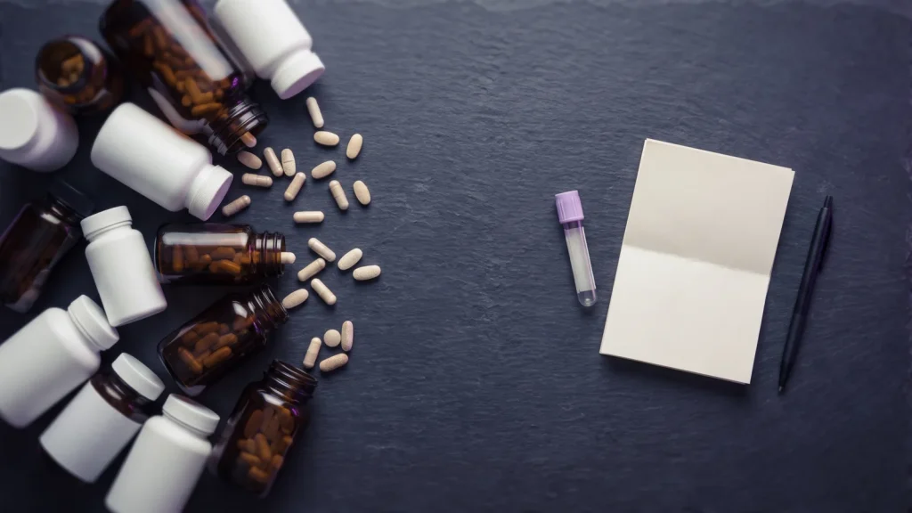 Unlabeled supplement bottles spilling capsules across dark slate, next to one blood collection vial and a blank test form