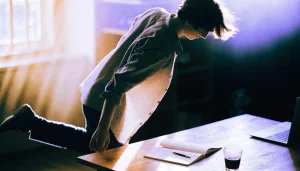 A person leaning over a desk in late afternoon light, pausing before stepping away, with an open notebook and pen beside a laptop.
